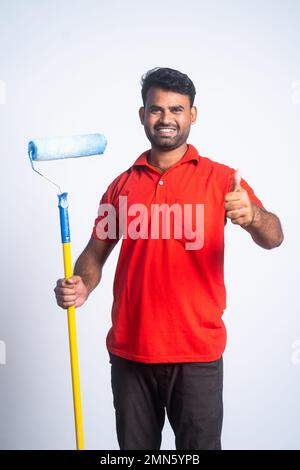 Vertical shot of smiling painter with paint roller brush showing thumbs up sign by looking camera - concept of professional service, employment and Stock Photo