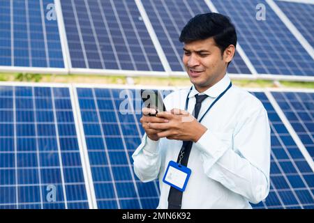 Young engineer busy using mobile phone in font of solar panel at power plant - concept of maintenance service, technology and renewable energy. Stock Photo