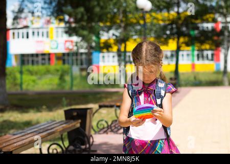 Little girl with a backpack and in a school uniform in the school yard plays pop it toy. Back to school, September 1. The pupil relaxes after lessons. Stock Photo