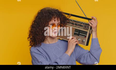 Curly haired woman using retro tape record player to listen music, disco dancing of favorite track, having fun, entertaining, fan of vintage technologies. Young teen girl isolated on yellow background Stock Photo