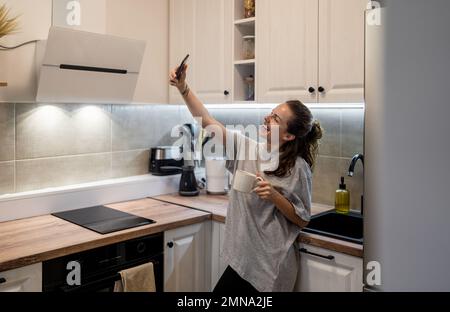 Woman photo messaging using her smart phone taking selfie while standing in kitchen at home in the morning and drinking coffee. Stock Photo