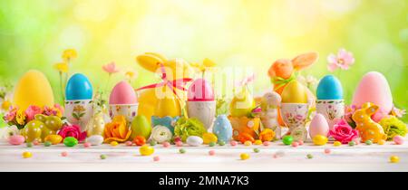 Easter concept with colorful decorated eggs in egg cups, rabbits and flowers on white wooden table on spring nature background. Easter background with Stock Photo