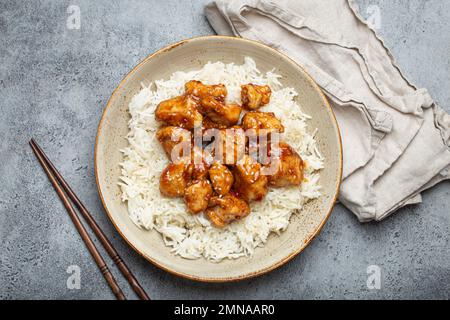 Chinese sweet and sour sticky chicken with sesame seeds and rice on ceramic plate with chopsticks top view, gray rustic stone background, traditional Stock Photo
