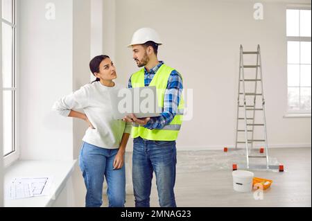 Attractive young woman is talking to construction foreman in white helmet, who demonstrates sequence of repairs in living room on laptop. Stock Photo
