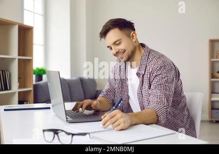 Happy student sitting at desk, using his modern laptop computer and taking notes Stock Photo