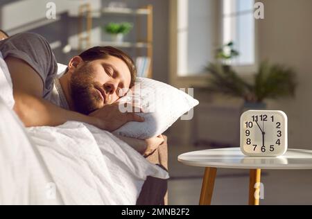 Handsome man sleeping on bed in his bedroom with alarm clock on his bedside table. Stock Photo