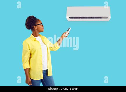 African american woman turns on air conditioner with a remote control on turquoise background. Stock Photo