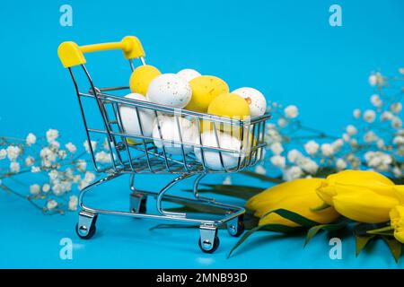 Easter white and yellow quail painted eggs in a shopping cart on a blue background. Easter Sale and shopping Stock Photo