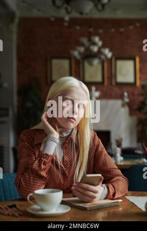 Restful albino girl with long blond hair looking at smartphone screen while chilling in cafe and searching for curious online video files Stock Photo