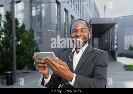 Portrait of young african american student in suit standing near campus wearing headphones. He holds a tablet in his hands, talks on a video call, listens to music, looks at the camera, smiles. Stock Photo