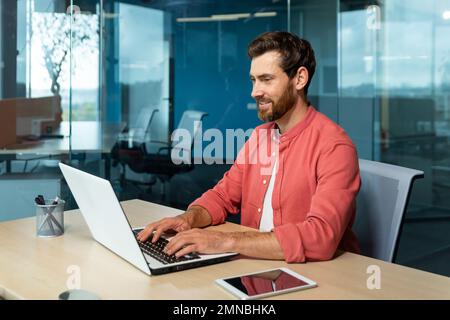 Distance Learning. Portrait of a young male student in a red shirt. Sitting at the desk in the office, studying online on a laptop. Writes a diploma, a course, an abstract, a lecture. Stock Photo