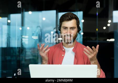 Distance Learning. Portrait of a young man teacher in a headset in a red shirt sits in the office at the table, works on a laptop online, teaches through a video call. Explains with his hands, smiles Stock Photo
