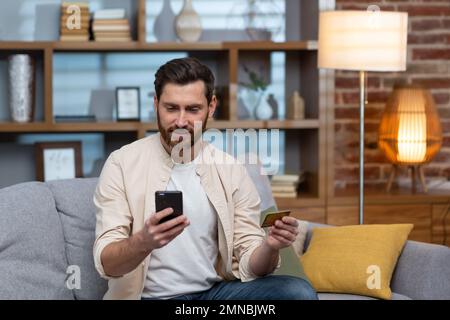 A young handsome man in a beige shirt is sitting on the sofa at home, using a phone and a credit card. Stock Photo