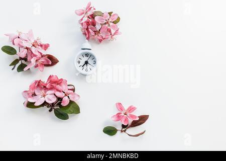 Small white clock with bell on white with fresh spring daisy flowers background. Spring forward, daylight saving time in March concept Stock Photo