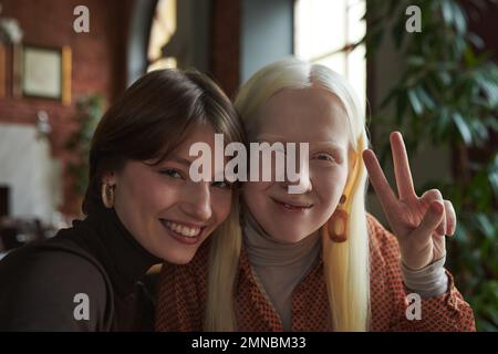 Young albino woman with long hair showing peace gesture while sitting next to her friend and both looking at camera in cafe Stock Photo