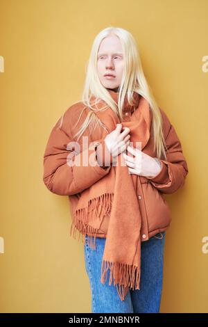 Long haired albino girl in blue jeans, brown jacket and scarf standing against yellow wall or background in isolation and looking aside Stock Photo