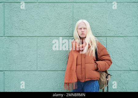 Adolescent albino girl in jeans, jacket and scarf standing by blue wall of building and looking at camera while posing in urban environment Stock Photo