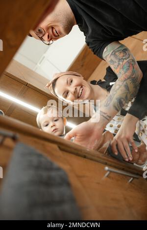 Joyful parents with little child in kitchen, from below view. Family portrait on stylish wooden kitchen furniture. Stock Photo