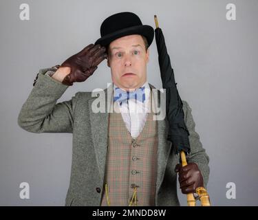 Portrait of British Character in Tweed Suit and Bowler Hat Holding Umbrella Like a Rifle and Saluting Fearfully. Concept of Vintage Vaudeville Actor Stock Photo