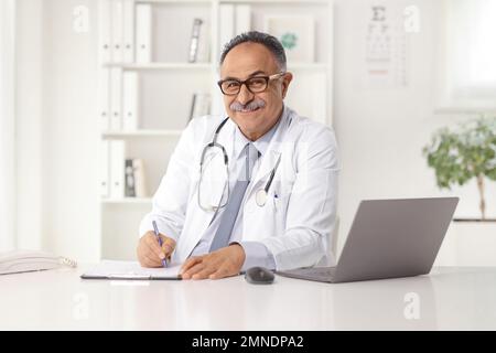 Senior male doctor writing a document in an office with a laptop on a desk, smiling and looking at camera Stock Photo