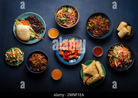 Set of assorted Chinese dishes on table: sweet and sour chicken on plate, fried spring rolls, noodles, salad, rice, steamed buns with bbq glazed Stock Photo