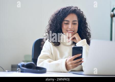 Young smiling latin business woman using phone in office sitting at desk. Stock Photo