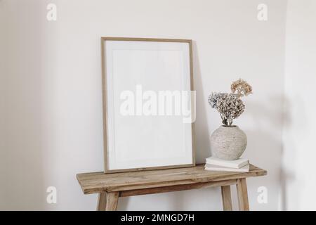 Neutral artistic still life. Blank picture frame mockup on old wooden bench in sunlight. Dry hydrangea flowers in textured vase. White wall background Stock Photo