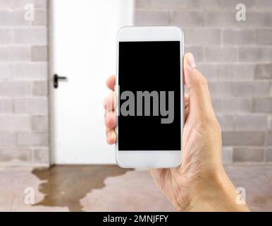 A hand holding up a smart phone with water damage in basement caused by sewer backflow due to clogged sanitary drain background Stock Photo