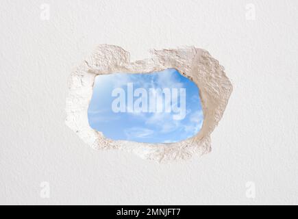 White wall with a hole that is observed the exterior of the building with clouds in the sky Stock Photo