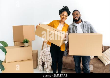 Moving to a new apartment. Happy newlyweds bought their first home, stand in the living room among the boxes, hold cardboard boxes in their hands, prepare to unpack things, look at the camera, smile Stock Photo