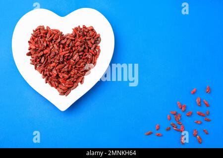 Dried goji berries in heart shaped plate on blue background, flat lay Stock Photo