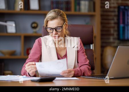 Portrait of adult businesswoman wearing eyeglasses working at office. Female manager doing paperwork, analyzing financial report, holding documents Stock Photo