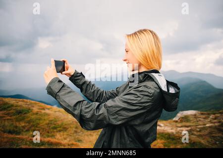 Casual young blonde hike woman taking pictures in his smartphone iPhone against a backdrop of mountains range during a thunderstorm cloud sky Stock Photo