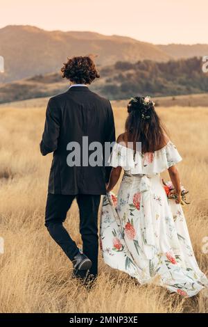 Newlywed couple walks through the countryside with bouquet in hand with the mountains in the background. Wedding Stock Photo