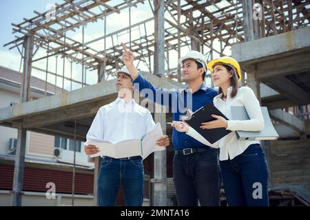 Asian Architector and engineers team with draft plan of building and tablet talking constructing site. Construction manager and engineer working on bu Stock Photo