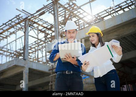 Asian Architector and engineers team with draft plan of building and tablet talking constructing site. Construction manager and engineer working on bu Stock Photo