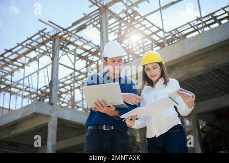 Asian Architector and engineers team with draft plan of building and tablet talking constructing site. Construction manager and engineer working on bu Stock Photo