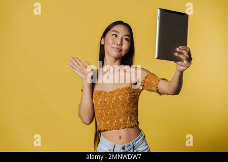 Young asian woman gesturing and using tablet computer isolated over yellow background Stock Photo