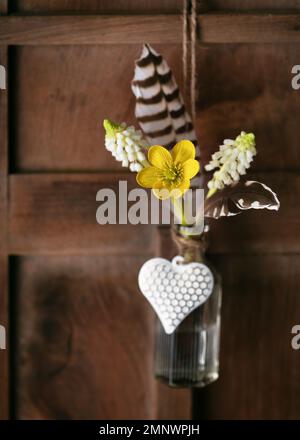 Floral decoration in the form of mini-vase and bouquet of yellow winter aconite wildflower, white grape hyacinth flowers, feathers and heart hanging Stock Photo