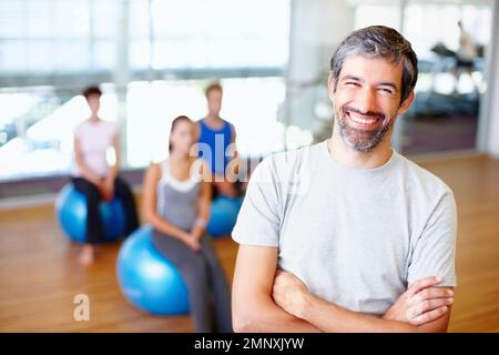 Confident male trainer smiling. Portrait of smiling male gym trainer with people practicing yoga in background. Stock Photo