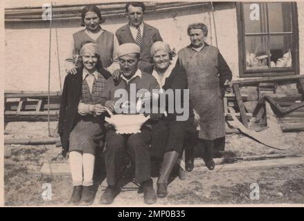 vintage moment / vintage funny moment / vintage photograph / power of the moment / magic moments / vintage eating / eating / vintage cooking / one man show from the past. A piping ( smoking) man is peeling potato for the lunch and two ladies are giving a help to him and behind the people give to him mental support :D funny moment from the 1930s. Stock Photo