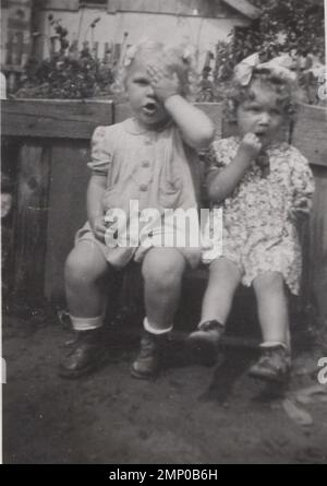 vintage moment / vintage funny moment / vintage photograph / power of the moment / magic moments / vintage photo about two little girls whose are making fun for the camera at the 1920s. One of them is hiding one of her eyes the other little girl is eating possibly cherry the fruit what they are holding in them hands. Stock Photo