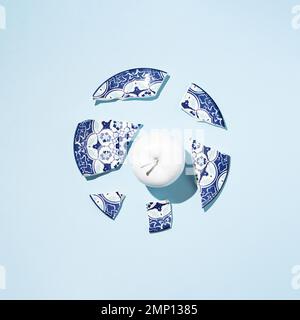 Fresh apple colored in white paint and broken china porcelain plate on blue background. Stock Photo
