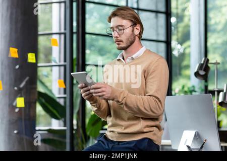 Young successful thinking programmer inside loft style office uses tablet computer, young blond man in casual clothes reads news online, serious businessman browses internet pages. Stock Photo