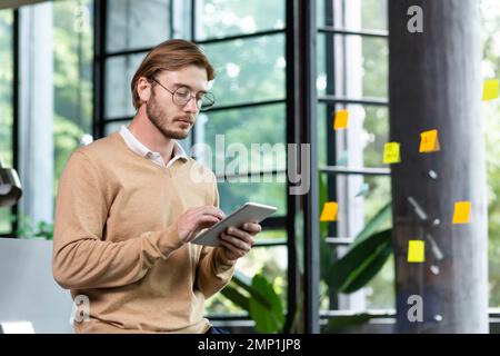 Young successful thinking programmer inside loft style office uses tablet computer, young blond man in casual clothes reads news online, serious businessman browses internet pages. Stock Photo