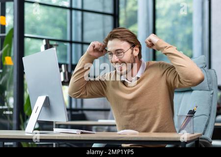 Young man in office celebrating victory and successful achievement results, businessman holding hands in triumph gesture and reading good news from computer , blond programmer in casual clothes. Stock Photo