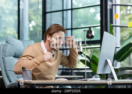 Young man in office celebrating victory and successful achievement results, businessman holding hands in triumph gesture and reading good news from computer , blond programmer in casual clothes. Stock Photo
