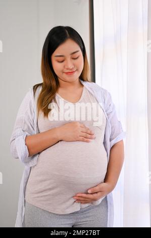 Relaxed and happy Asian pregnant woman in casual clothes feeling her baby, touching her belly with love and care. parenting and motherhood concept Stock Photo