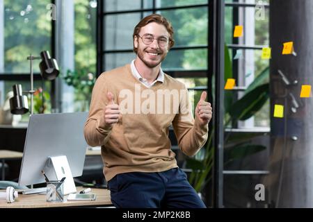 Portrait of happy blond man inside office, man in casual clothes smiling and looking at camera, young successful businessman showing thumbs up at camera, programmer at work with computer. Stock Photo