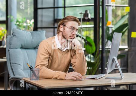 Frustrated and upset businessman reading news online from computer monitor, young blond man sad at workplace inside office in casual clothes unhappy with results. Stock Photo